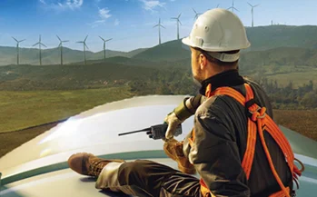Technician in hard hat and safety harness using a two-way radio atop a wind turbine